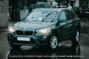 “Dark blue BMW SUV driving through a rainy city street with headlights on and windshield wipers active. Wet pavement reflects the lights, and pedestrians with umbrellas are visible in the background.”