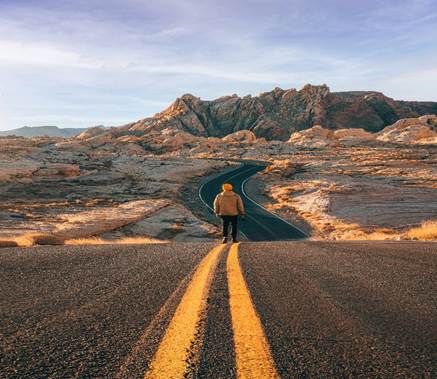 A person stands alone on a winding desert road at sunrise, facing the distant mountains, symbolizing life’s uncertain path and the constant change ahead.