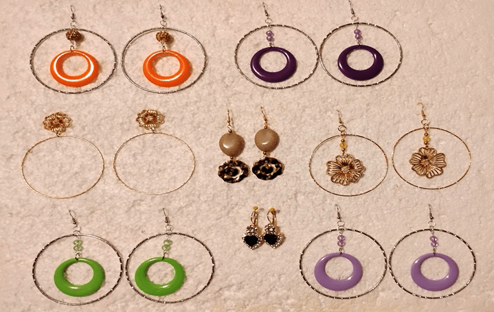 A neatly arranged collection of handmade earrings displayed on a light textured surface. The photo shows twelve unique pairs in bright colors and varied designs—large hoops with colorful pendants, floral motifs, heart shaped stones, and beadwork—showcasing the one of a kind style of Penny Pinching Jewelry.