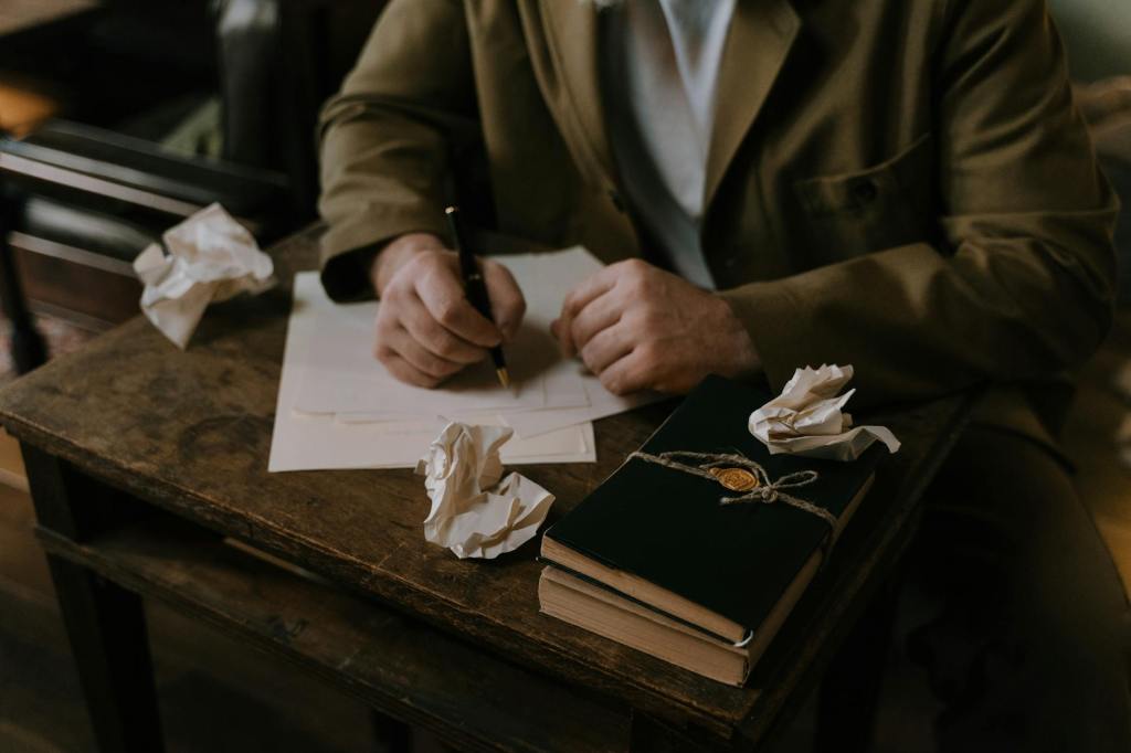 A person sits at a wooden desk writing on paper, surrounded by crumpled drafts and two closed books, creating a warm, vintage scene of persistence and creativity.