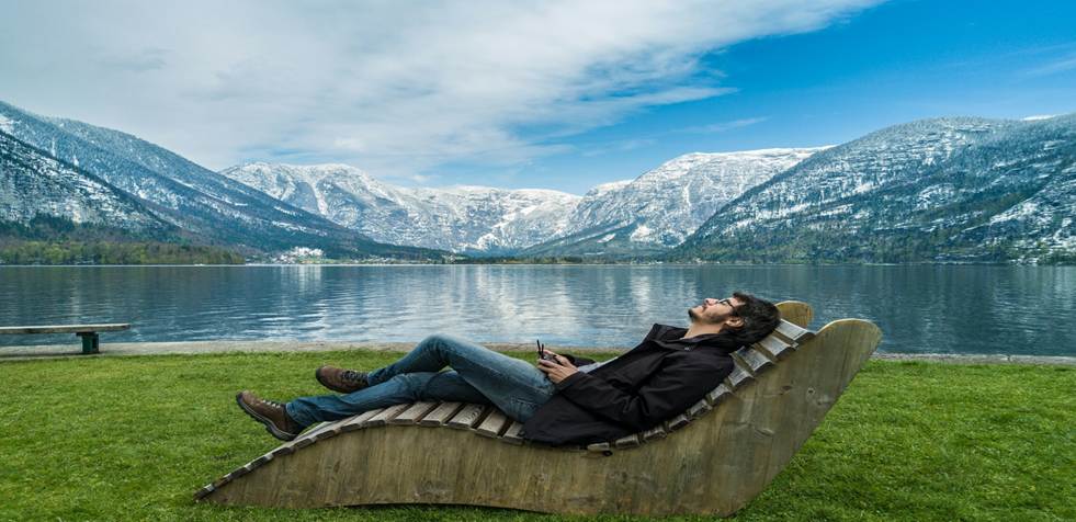 A man reclines in a curved wooden lounge chair beside a calm lake, mountains rising in the distance. The scene is peaceful and quiet, suggesting rest, reflection, and stress relief.