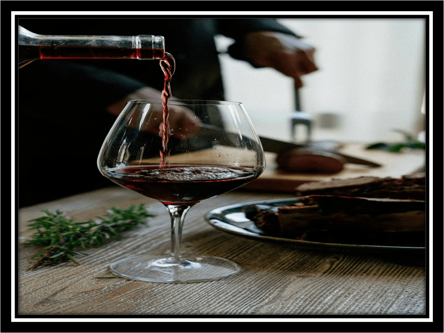 A moody photo of red wine being poured into a glass, framed in black, with candlelight and rustic tones.