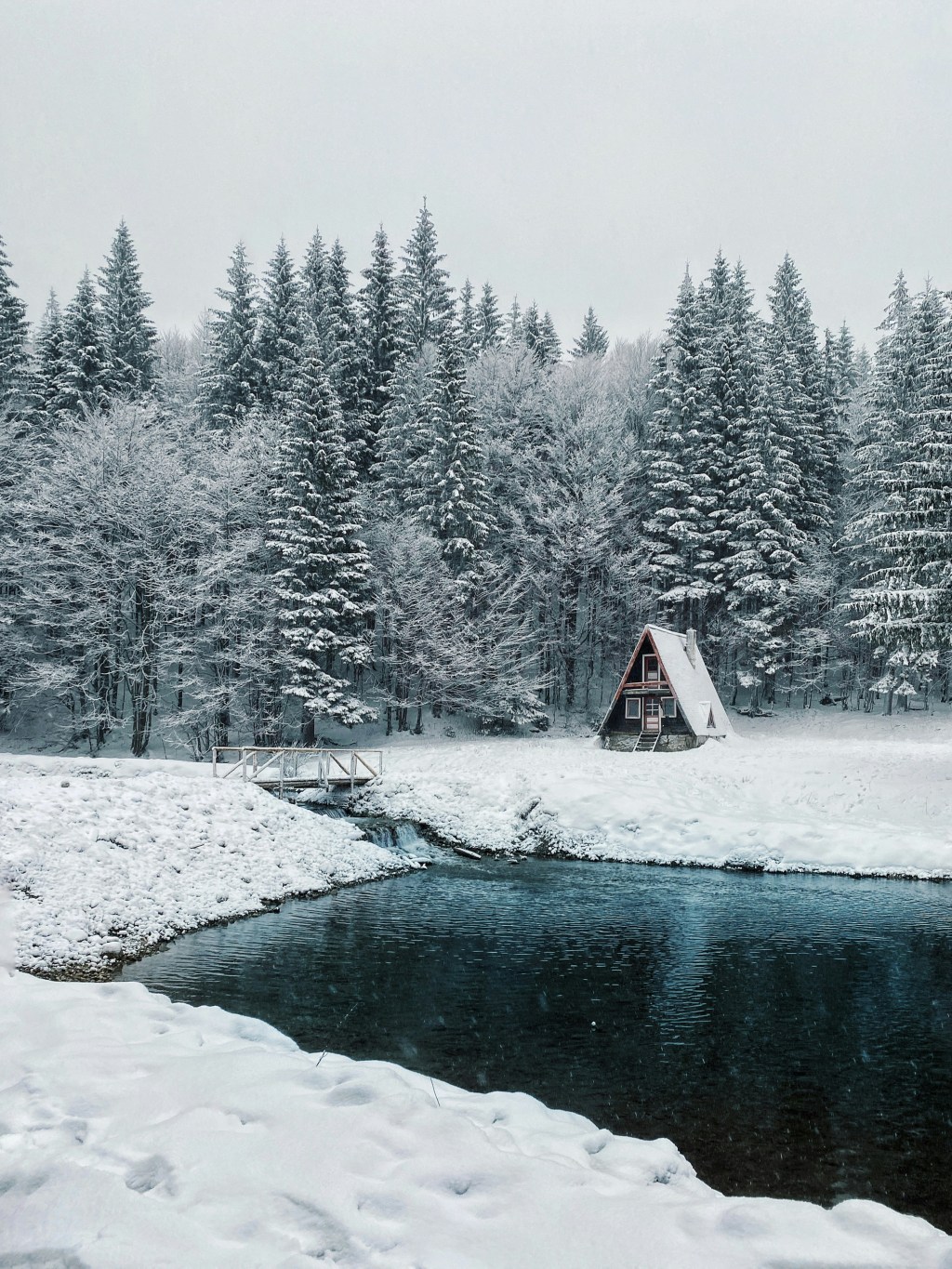 Snow‑covered forest with a small A‑frame cabin beside a quiet pond, capturing the calm beauty of winter described in Debra A Winter (Richter)’s haiku Heavenly Bliss.